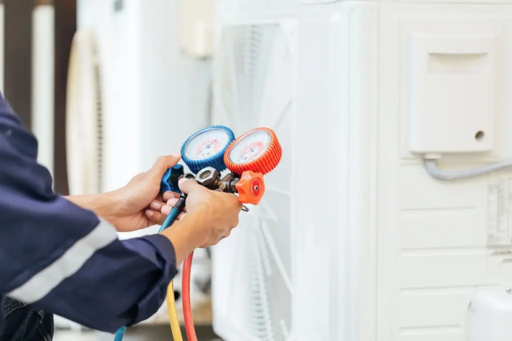 Technician's hands holding blue and red manifold gauges with colored hoses beside a white outdoor HVAC unit.
