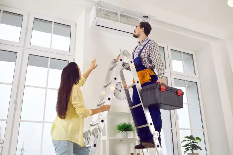 HVAC technician on a ladder holding a toolbox while inspecting a wall-mounted mini split unit as a homeowner watches.