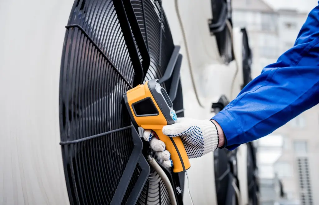 Gloved hand holding an infrared thermometer against the fan grille of a large commercial HVAC condenser unit.