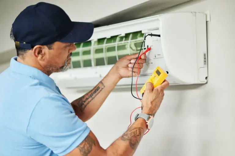 Technician in a cap uses a yellow multimeter to test wiring on a wall-mounted ductless mini split indoor unit.
