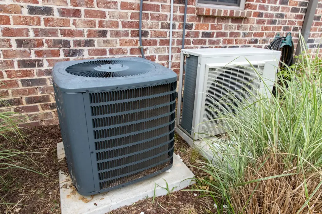 Gray central air conditioning condenser and white ductless heat pump unit on concrete pads beside a brick house exterior.