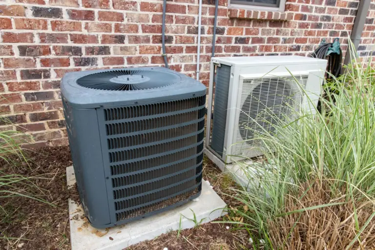 Gray central air conditioning condenser and white ductless heat pump unit on concrete pads beside a brick house exterior.