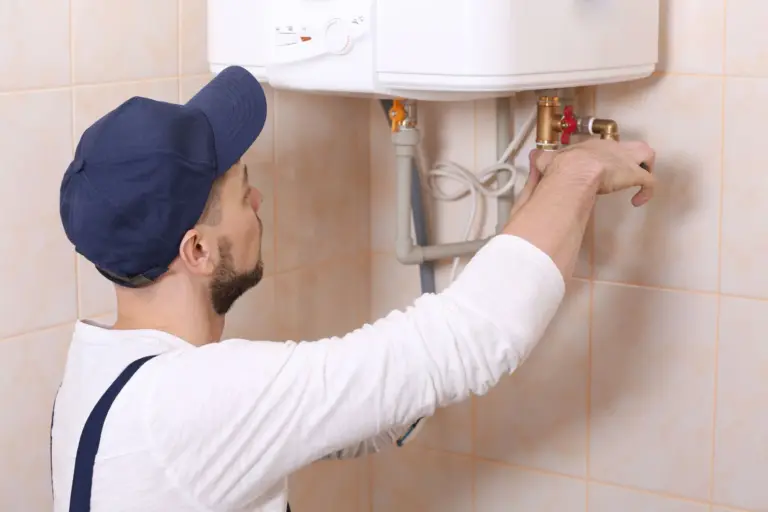 Technician in a blue cap and overalls adjusting a brass valve on a wall-mounted white water heater in a tiled room.