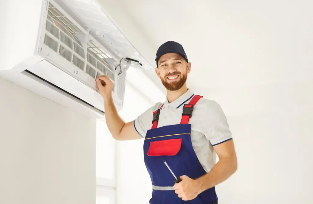 Smiling technician in cap and blue overalls holds a screwdriver while servicing an open wall-mounted mini split indoor unit.